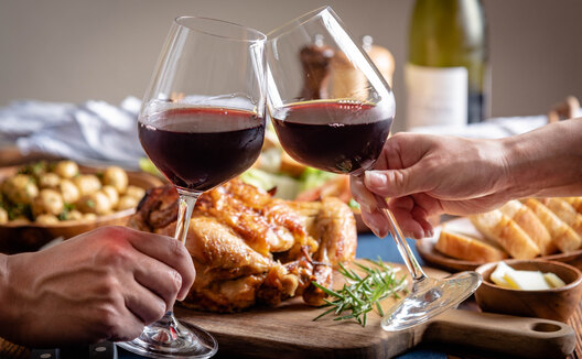 Two hands "cheersing" with glasses of red wine at Table with Thanksgiving dishes including turkey and potatoes